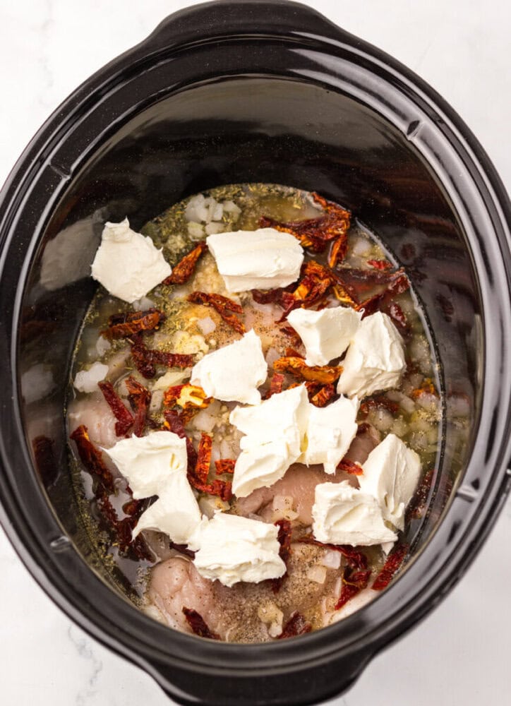 Chicken, sun-dried tomatoes, onions, and cream cheese in a slow cooker, ready to be cooked.