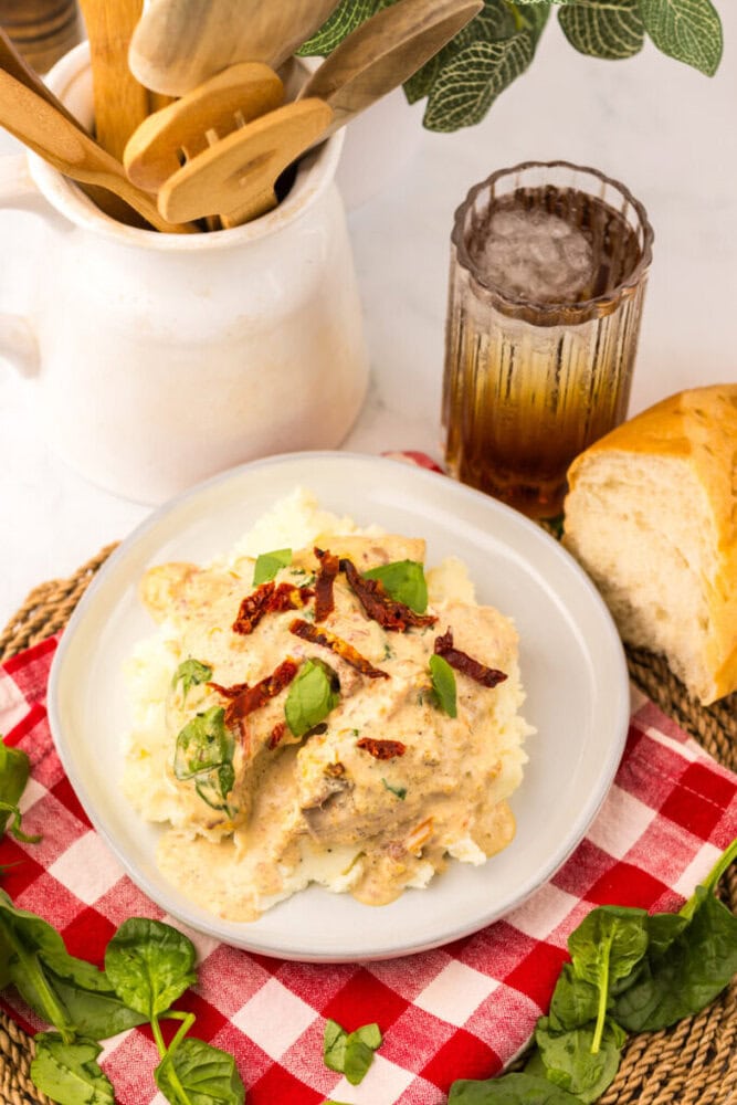 Plate of creamy chicken on mashed potatoes, garnished with herbs and sun-dried tomatoes, next to bread and a drink.