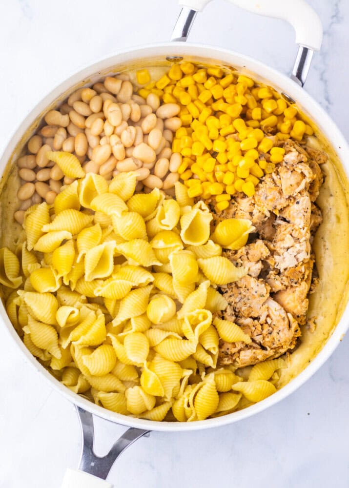 A white pot containing cooked pasta shells, white beans, corn, and a block of seasoned tofu on a white surface.