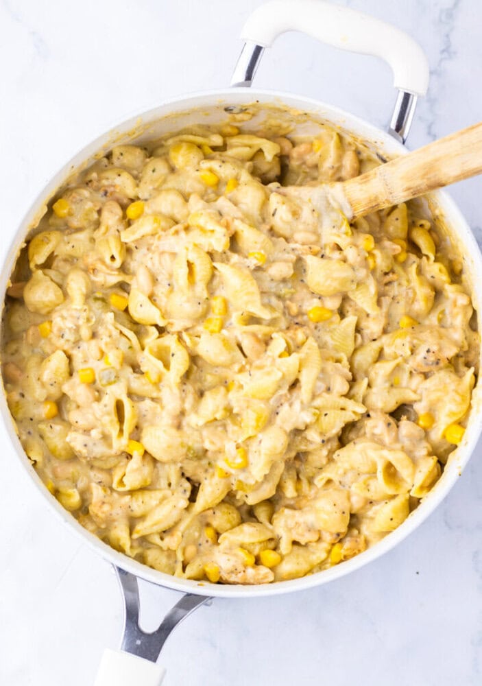 A white pot filled with creamy shell pasta mixed with corn and a wooden spoon resting inside.
