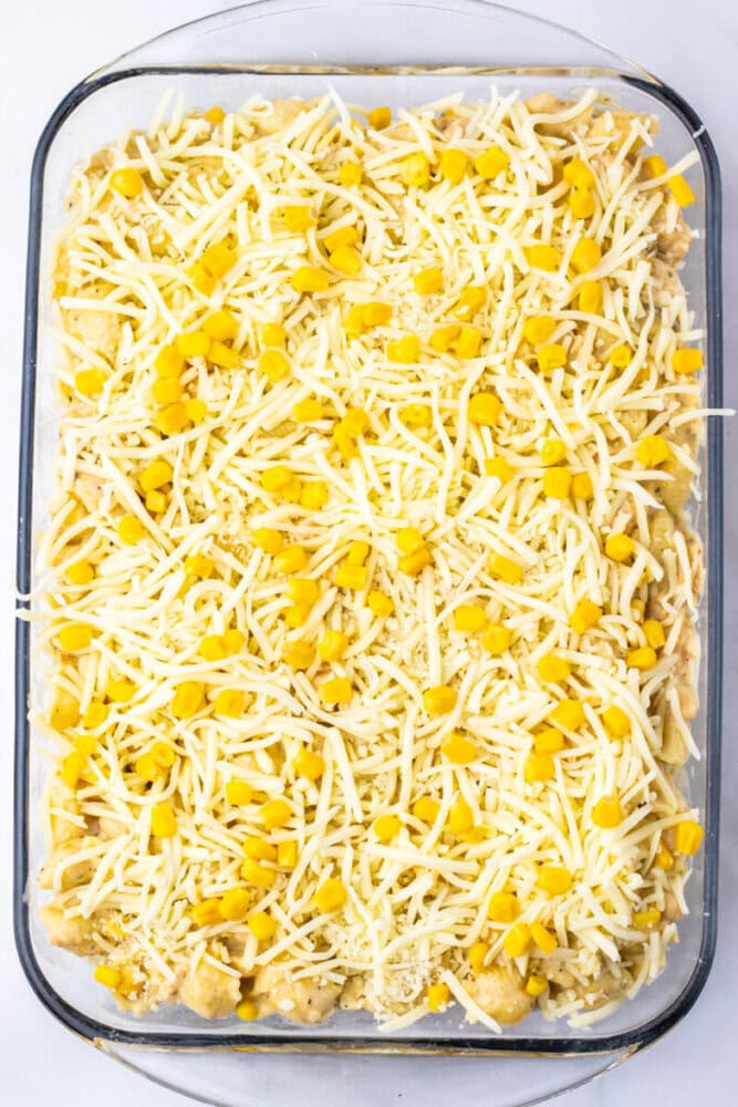 A glass baking dish filled with an unbaked casserole topped with shredded cheese and yellow corn kernels.