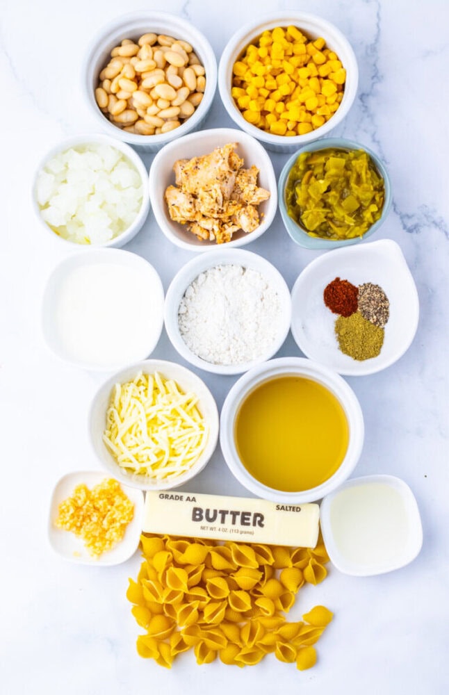 Assorted ingredients in small bowls including beans, corn, diced onion, green chiles, spices, shredded cheese, milk, flour, butter, oil, garlic, and uncooked shell pasta on a white surface.