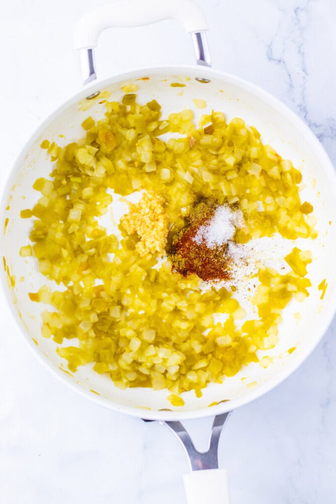 Diced onions and garlic are saut&eacute;ing in a white pan with added spices, including salt and paprika, on a marble countertop.