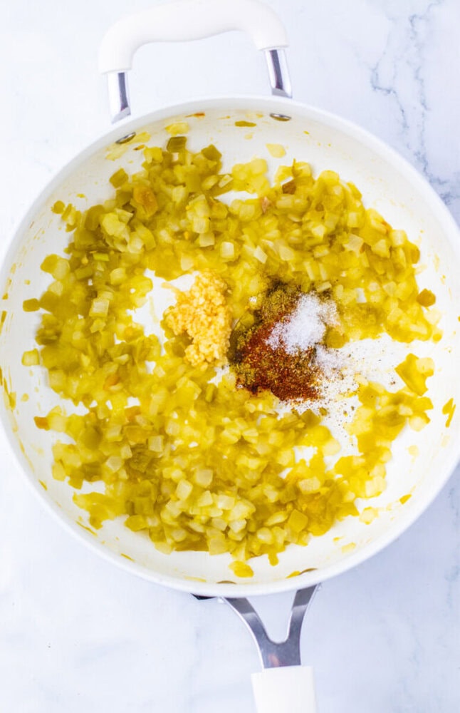 Chopped onions saut&eacute;ing in a white pan with minced garlic, spices, and salt added on top, on a marble surface.