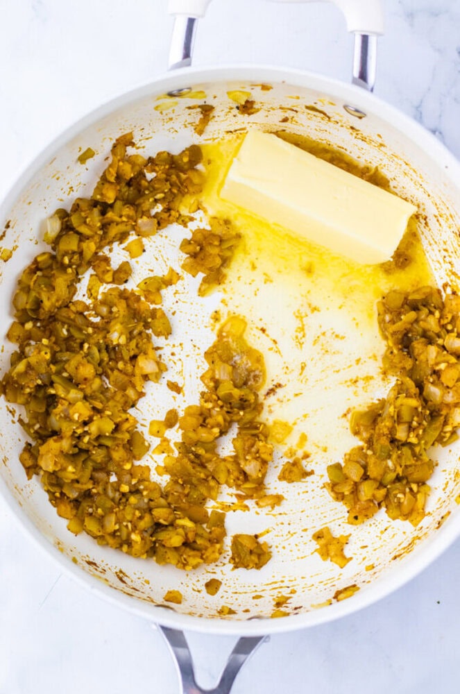 A white pan containing saut&eacute;ed chopped onions and spices, with a stick of butter melting on one side.