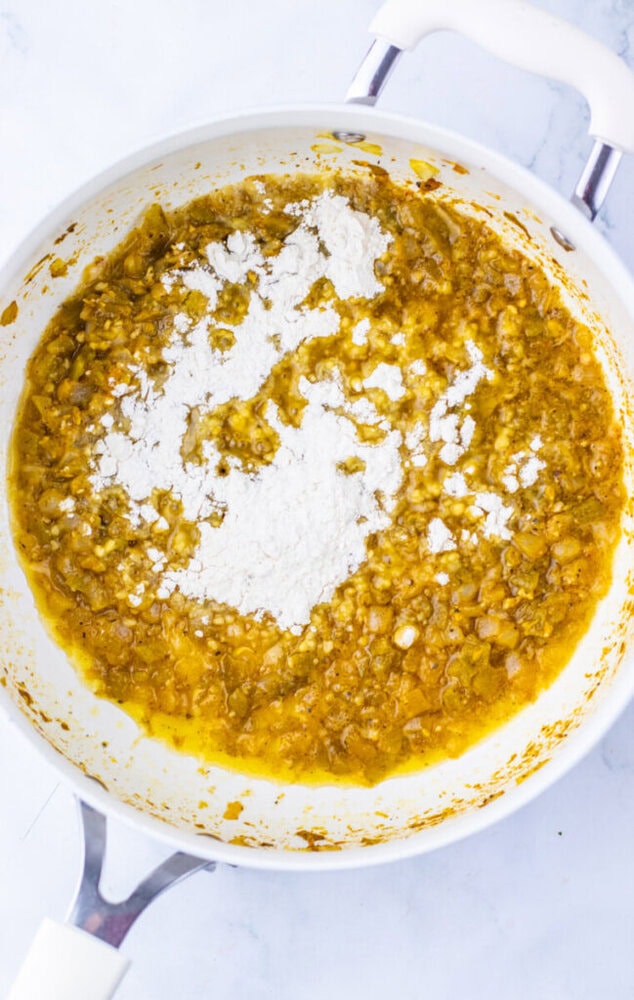 A white pan on a stove contains a yellowish mixture of saut&eacute;ed onions and spices with a mound of flour sprinkled on top.
