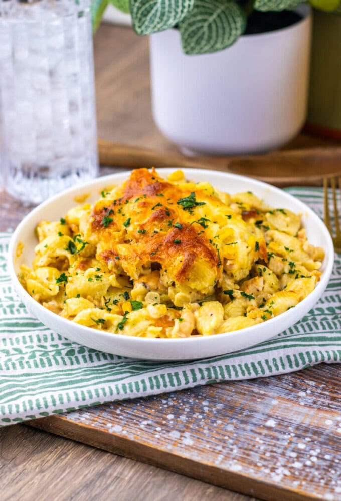 A bowl of baked macaroni and cheese topped with melted cheese and chopped herbs, placed on a patterned napkin beside a glass of water and a potted plant.