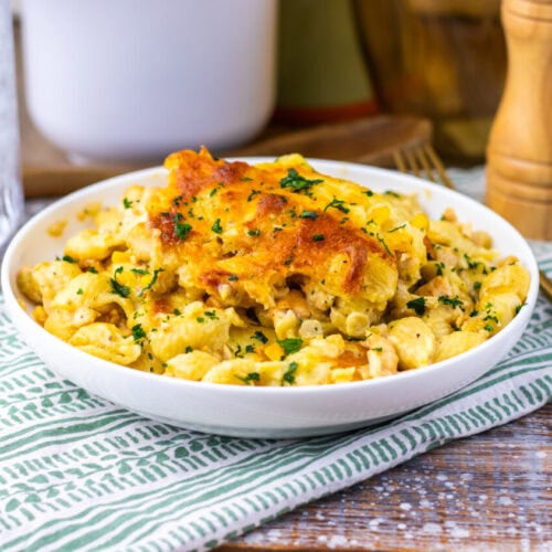 A bowl of baked macaroni and cheese with a crispy golden top, garnished with chopped herbs, sits on a striped cloth next to a glass of ice water and wooden salt and pepper grinders.