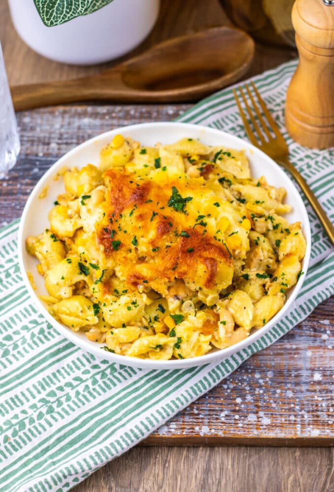 A bowl of baked macaroni and cheese garnished with chopped parsley sits on a cloth napkin, with a fork and pepper grinder nearby.