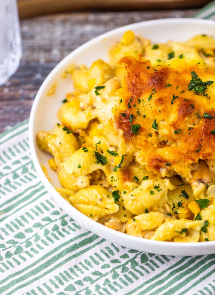 A close-up of baked macaroni and cheese with a golden, crispy top, garnished with chopped parsley, served in a white bowl on a green and white patterned cloth.