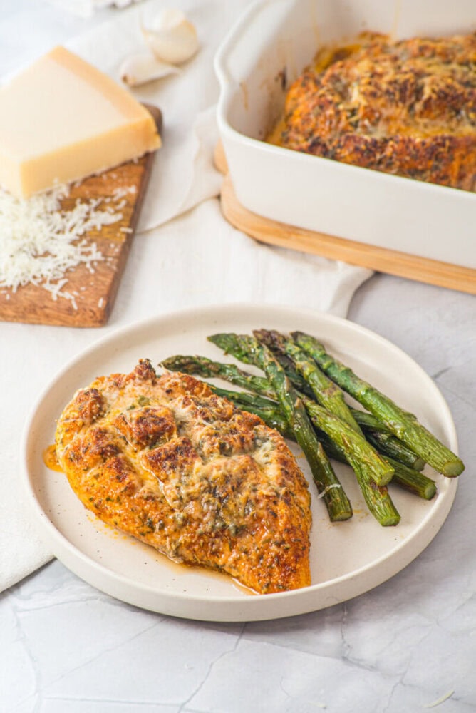 Baked chicken breast with herbs and roasted asparagus on a plate, with cheese and a baking dish in the background.