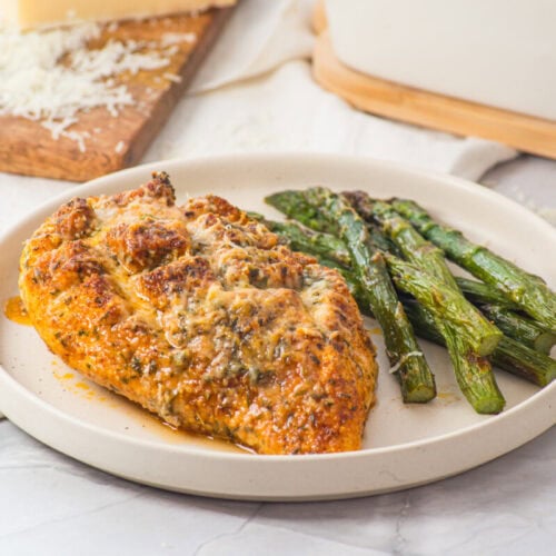 Seasoned baked chicken breast with melted cheese, served with roasted asparagus on a beige plate.