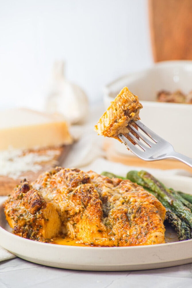 A fork holds a bite of cheesy baked chicken with herbs, served with roasted asparagus on a plate.