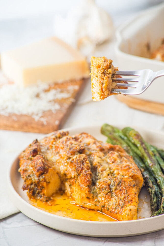 A fork holds a bite of cheesy baked chicken with roasted asparagus on a plate beside it.