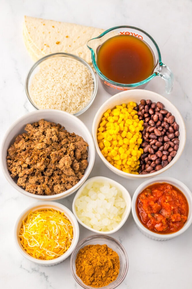 Ingredients for a taco dish: rice, broth, tortillas, ground meat, corn, black beans, onions, salsa, cheese, seasoning.