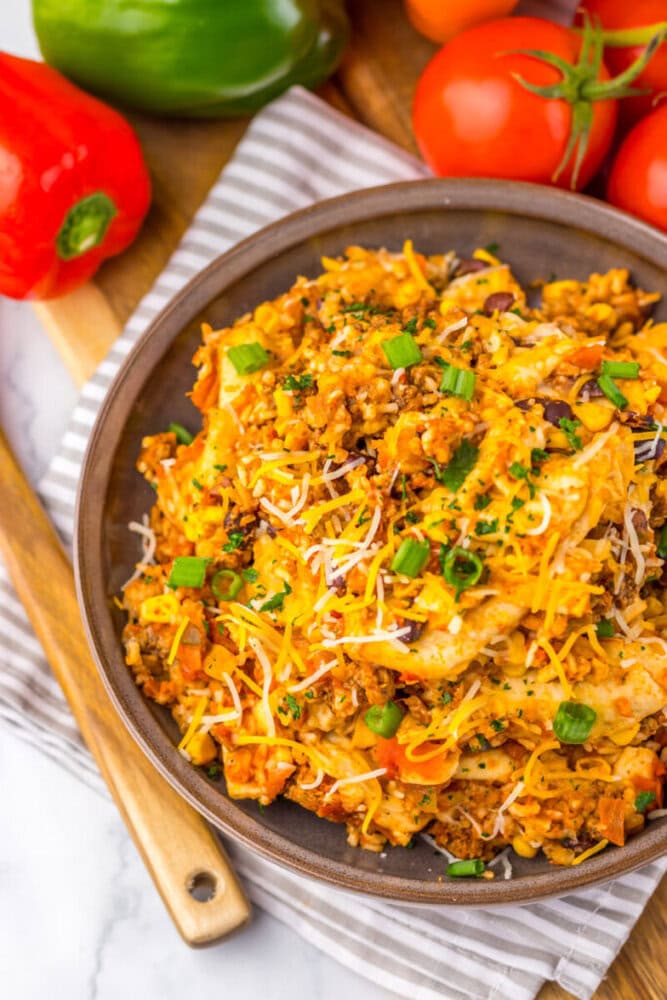 A bowl of cheesy ground beef and rice topped with chopped green onions, surrounded by fresh vegetables.