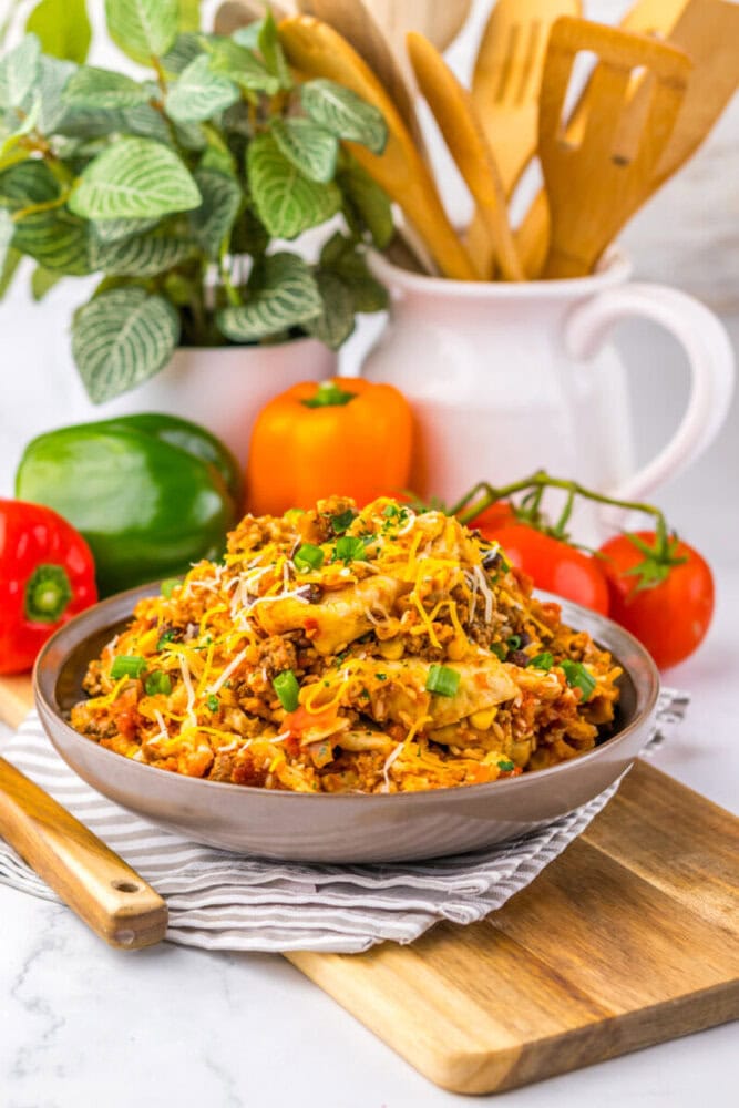 A bowl of cheesy chicken and rice topped with herbs, surrounded by fresh peppers and tomatoes on a kitchen counter.