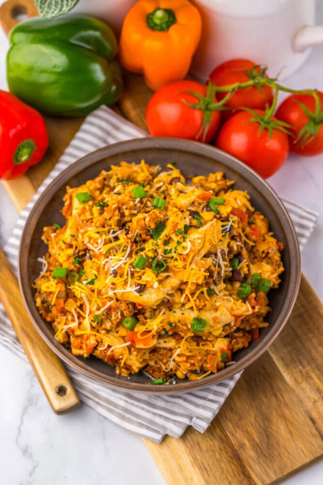 A bowl of cheesy rice and ground meat with vegetables, topped with green onions, next to fresh peppers and tomatoes.