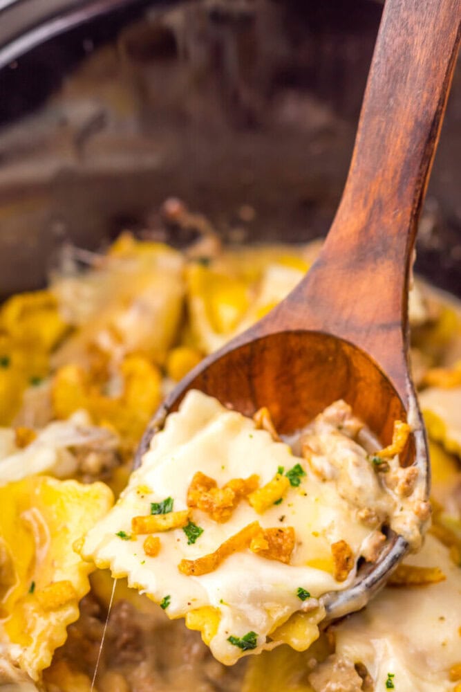 Close-up of a wooden spoon lifting cheesy ravioli with ground meat and crispy onions from a creamy dish.