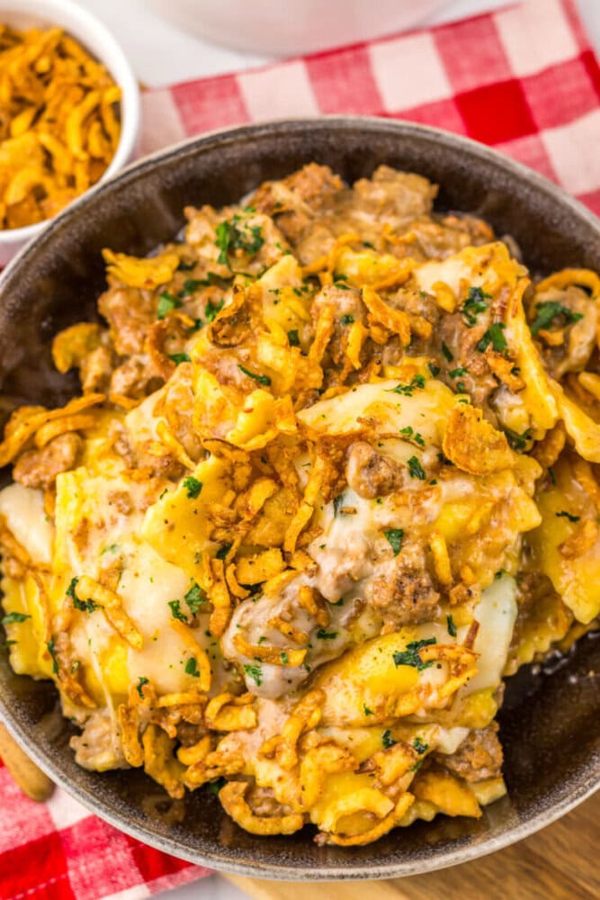 A bowl of cheesy ravioli casserole topped with ground beef, crispy fried onions, and parsley on a red checkered tablecloth.