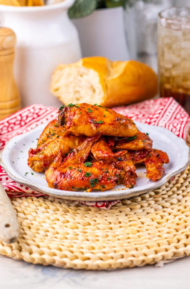 A plate of glazed chicken wings garnished with herbs, with bread and a drink in the background.