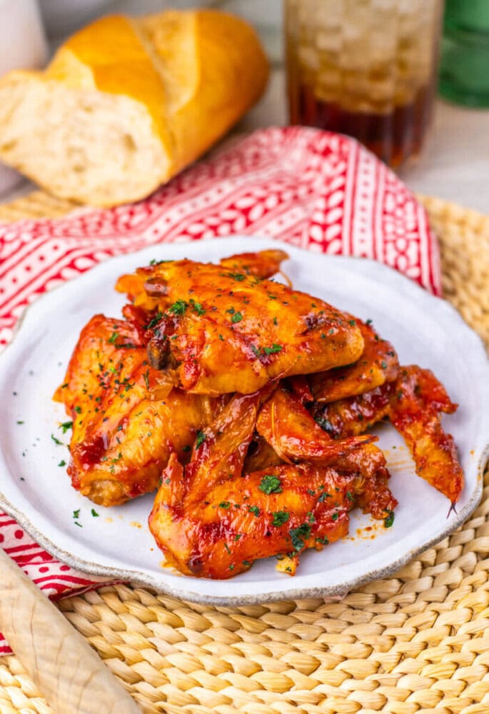 A plate of glazed chicken wings garnished with herbs, with bread and a drink in the background.