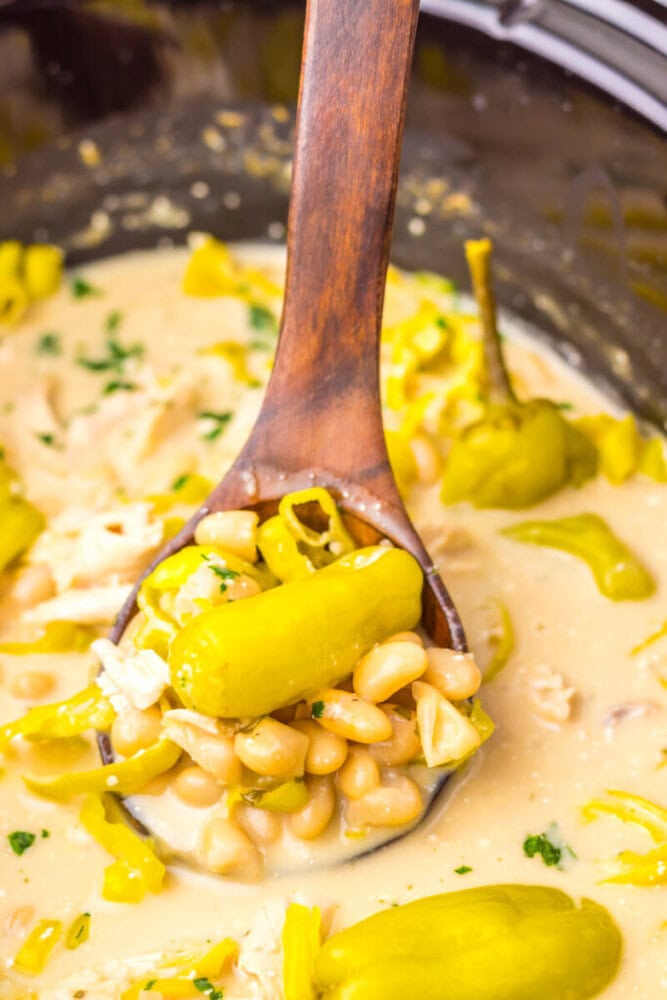 A wooden spoon scoops creamy white bean chili with shredded chicken and whole yellow peppers.