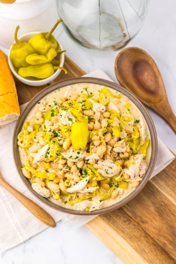 A bowl of white bean chicken chili topped with sliced pepperoncini and herbs, beside bread and a wooden spoon.