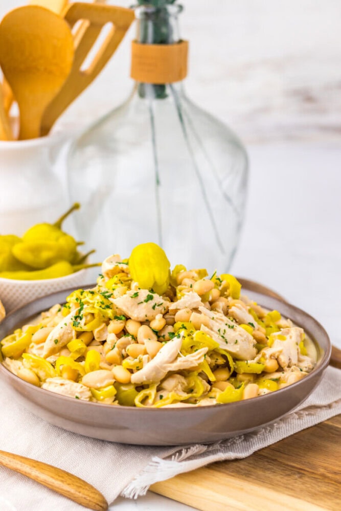 A bowl of shredded chicken, white beans, and peppers garnished with herbs and a whole pepper on top.