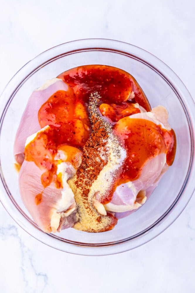 Raw chicken breasts in a glass bowl topped with spices, garlic, and a reddish sauce on a white surface.