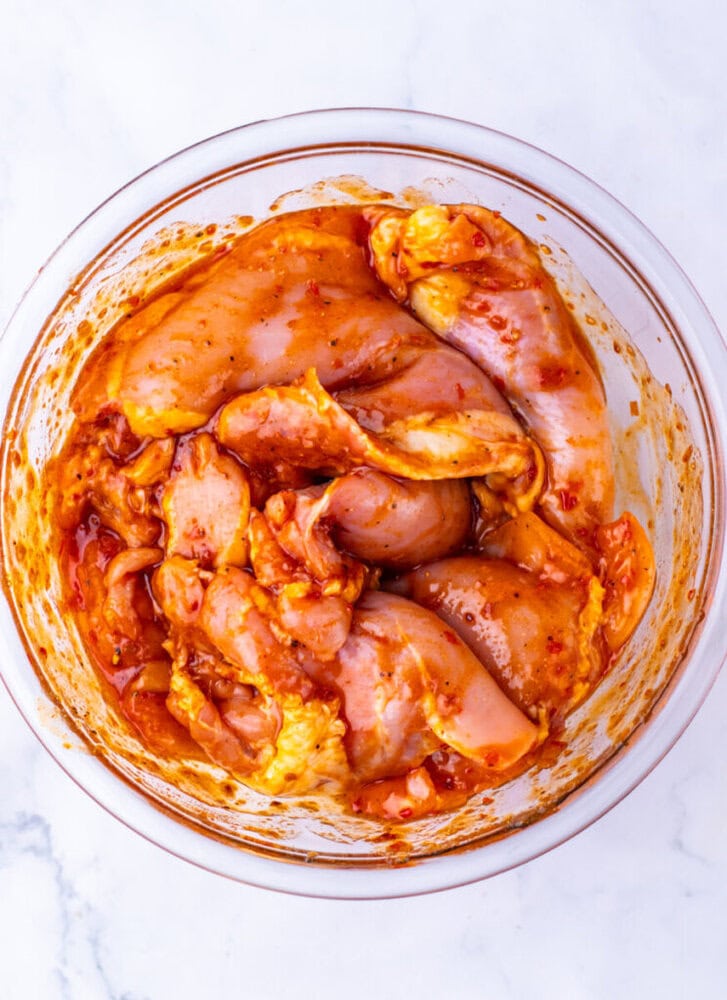 Raw chicken pieces marinated in a reddish spice mixture in a glass bowl on a marble surface.