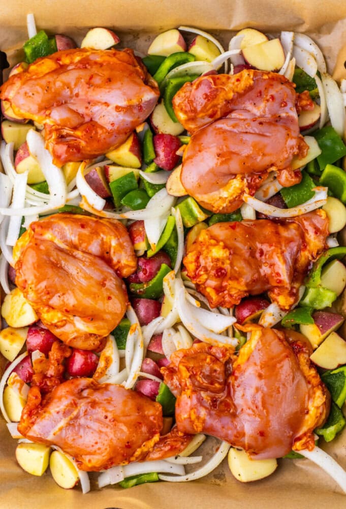 Raw marinated chicken thighs on a bed of chopped potatoes, onions, and bell peppers in a baking dish.
