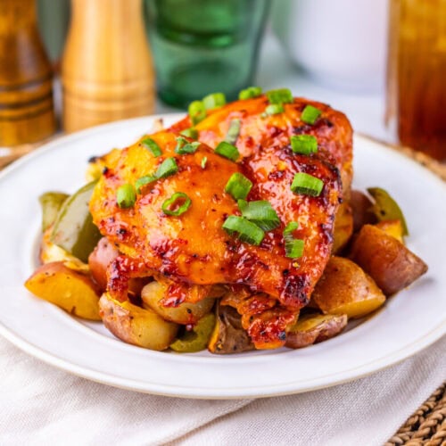 Baked chicken thighs with green onions on roasted potatoes and peppers, served on a white plate.