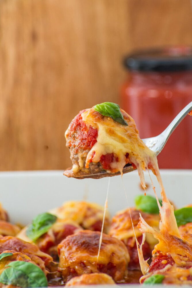 A cheesy meatball with tomato sauce and basil is lifted from a baking dish with a fork.