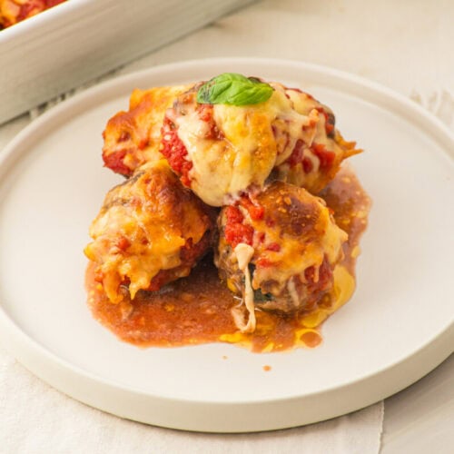 Three baked eggplant rolls topped with melted cheese and tomato sauce on a white plate, garnished with basil.
