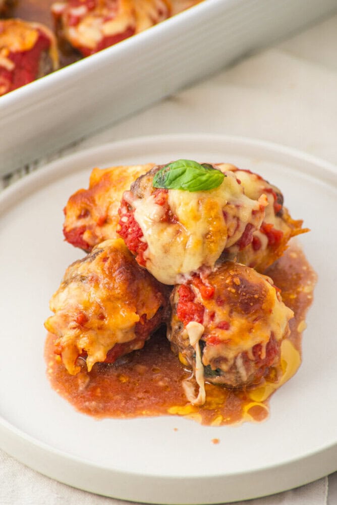 Three cheesy baked meatballs with tomato sauce on a white plate, garnished with a basil leaf.