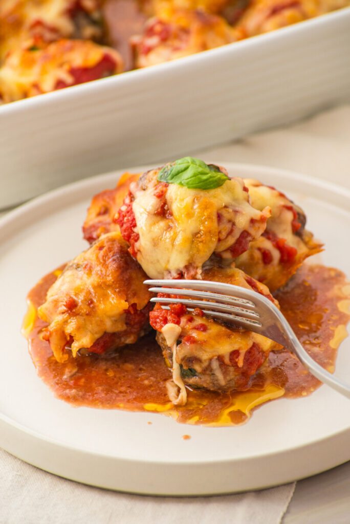 Three cheesy, baked meatballs topped with tomato sauce and basil on a white plate, with a fork and sauce.
