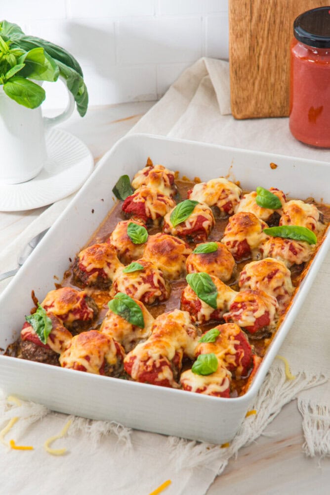 A white baking dish of cheesy baked meatballs topped with tomato sauce and fresh basil leaves.