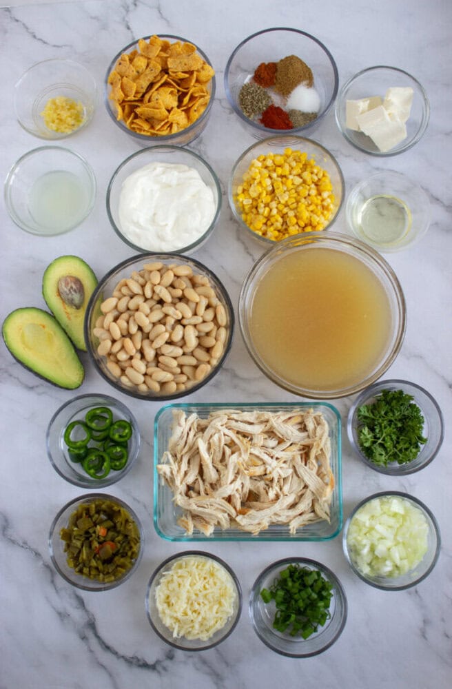 Ingredients for white chicken chili in bowls, including beans, corn, shredded chicken, avocado, cheese, and spices.