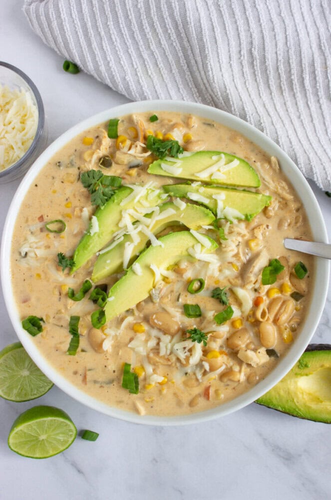 A bowl of creamy white chili topped with avocado, shredded cheese, and green onions, with lime slices beside it.