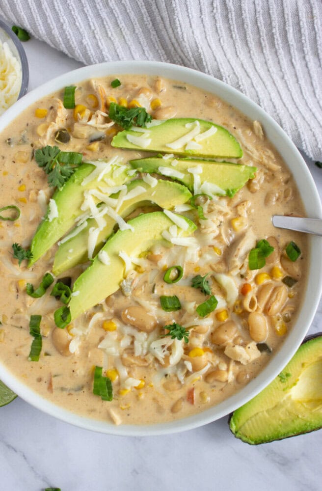Bowl of creamy white chicken chili topped with sliced avocado, shredded cheese, and chopped green onions.