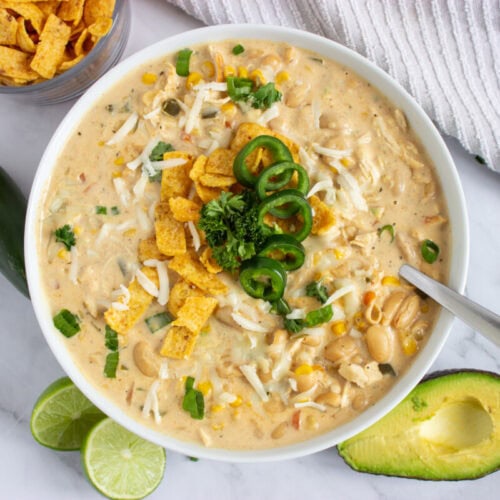 A bowl of creamy chili topped with cheese, jalape&ntilde;os, corn chips, and herbs, with avocado and lime on the side.