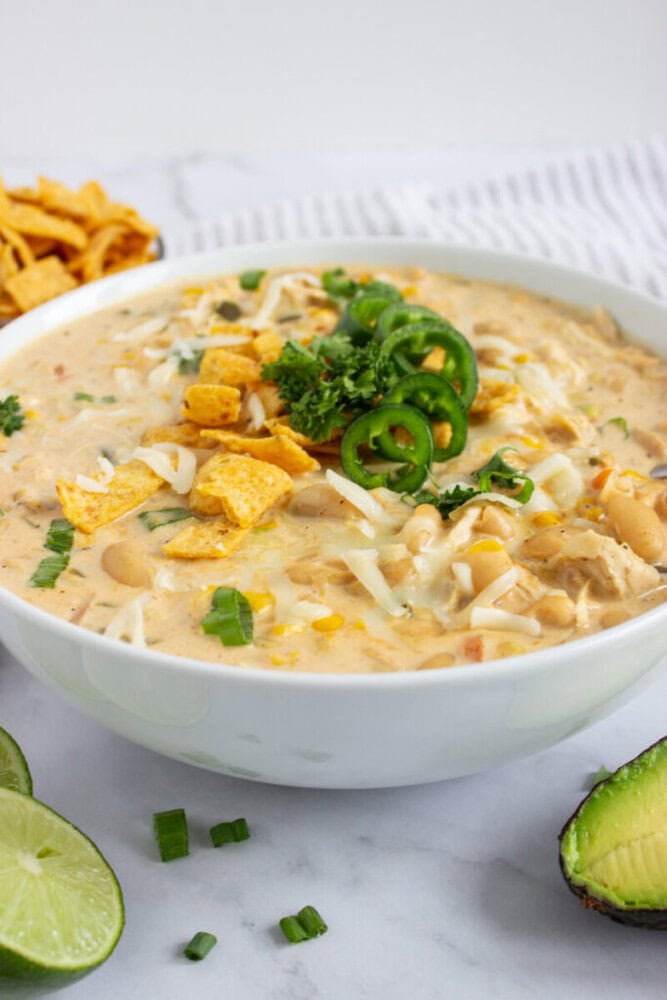 A bowl of creamy white chili topped with Fritos, jalape&ntilde;os, shredded cheese, and fresh herbs.
