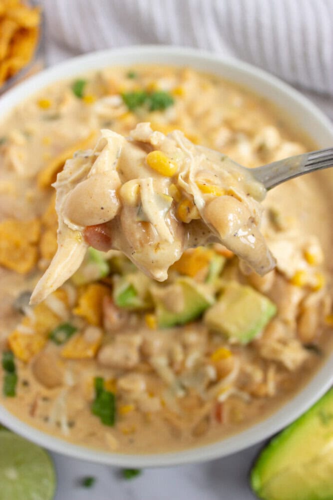 A spoonful of creamy white chili with chicken, beans, corn, and avocado held above a bowl.