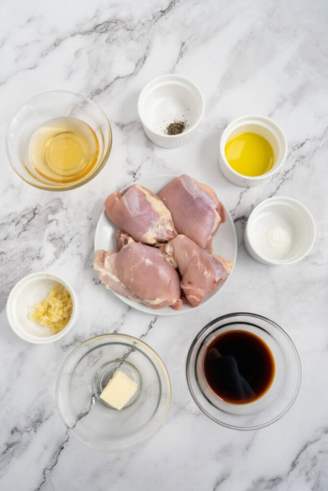 Raw chicken thighs on a plate, surrounded by bowls of oil, soy sauce, garlic, butter, and seasonings on marble.