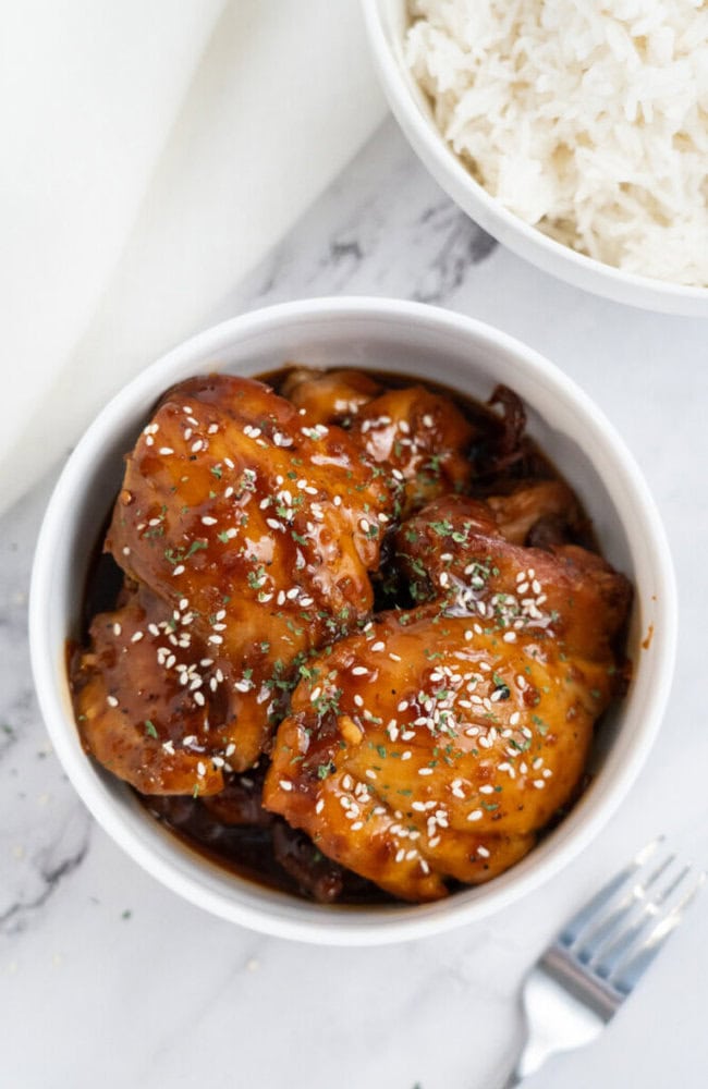 Bowl of glazed chicken topped with sesame seeds and herbs, next to a bowl of white rice on a marble surface.