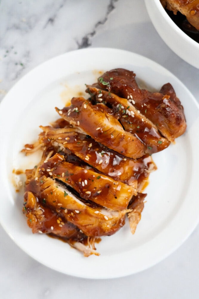 Sliced teriyaki chicken breast topped with sesame seeds and sauce on a white plate.