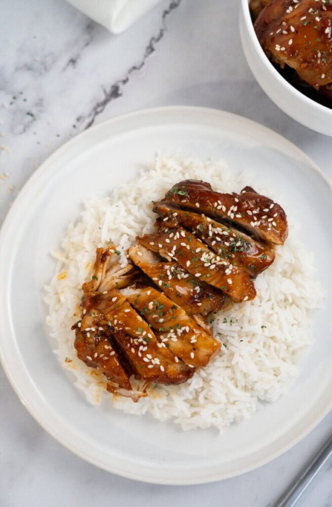 Sliced teriyaki chicken with sesame seeds served over white rice on a white plate.