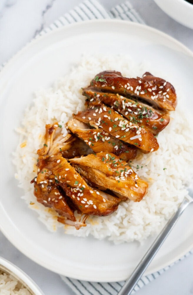Sliced teriyaki chicken garnished with sesame seeds served over white rice on a white plate.