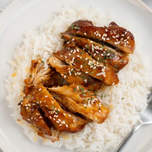 Sliced teriyaki chicken with sesame seeds served on white rice, on a white plate with a fork.